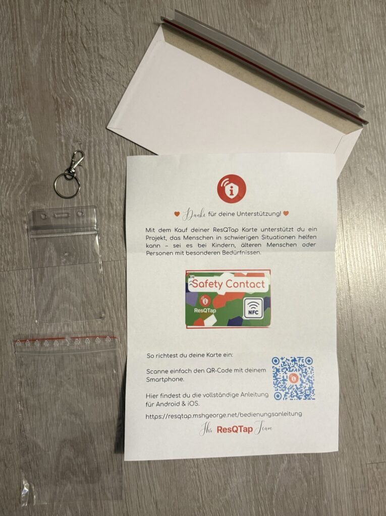 Ein Briefumschlag mit dem Begrüßungsschreiben, in der mitte die Saftey Contact Card, ein Karabinerhaken, Schlüsselring und das Etui.